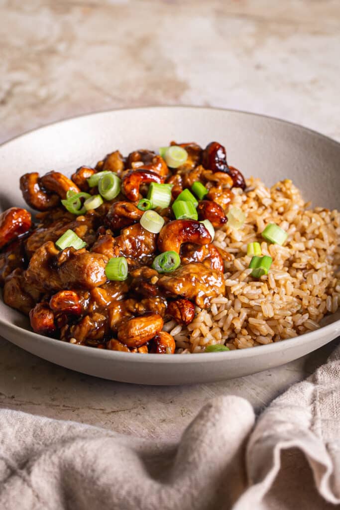 a close up horizontal image of brown rice in a bowl topped with vegan sticky cashew chicken and garnished with sliced green onion