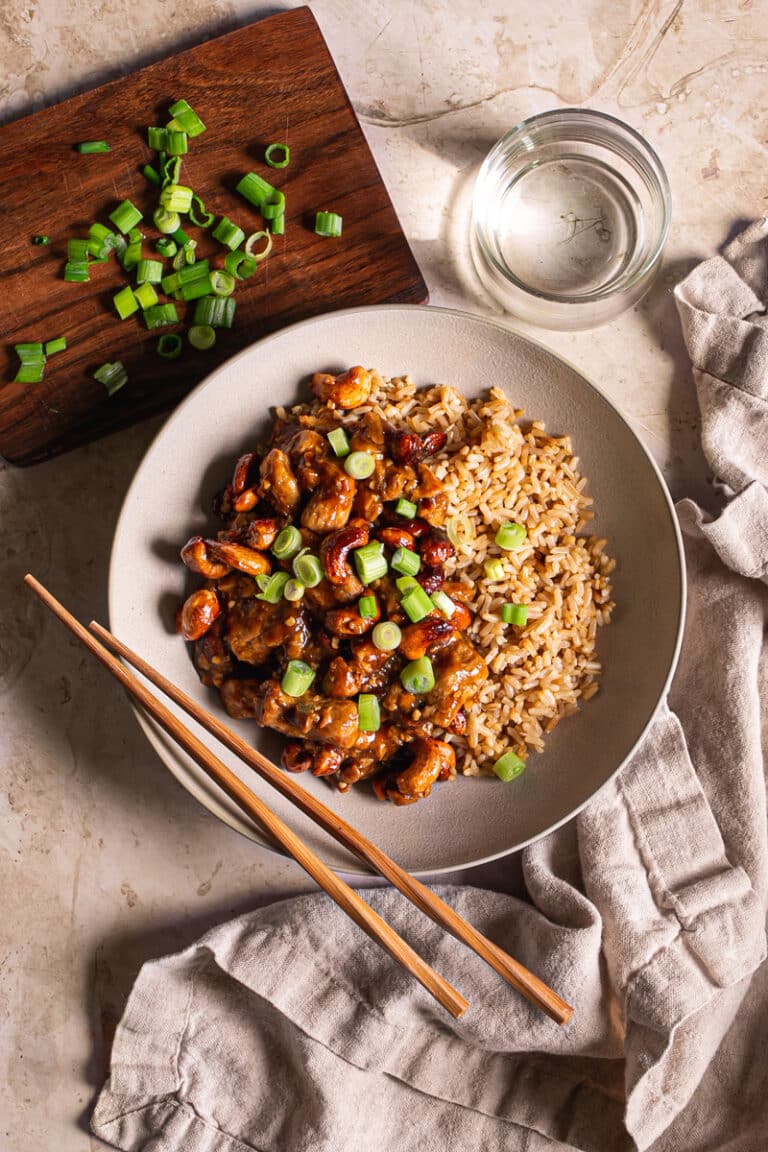 a bowl of sticky vegan cashew chicken served over brown rice with chopsticks resting on the bowl, a glass of water on the side, a small wooden cutting board with sliced green onions on it.