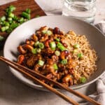3/4 shot of a bowl of sticky vegan cashew chicken served over brown rice with chopsticks resting on the bowl, a glass of water on the side, a small wooden cutting board with sliced green onions on it.