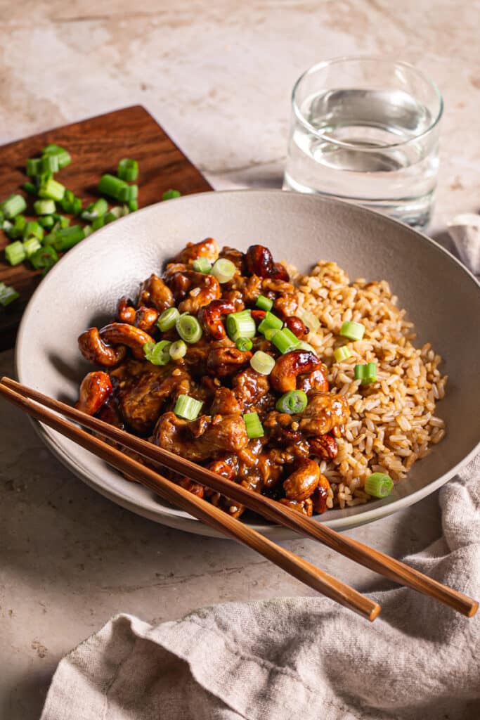 3/4 shot of a bowl of sticky vegan cashew chicken served over brown rice with chopsticks resting on the bowl, a glass of water on the side, a small wooden cutting board with sliced green onions on it.