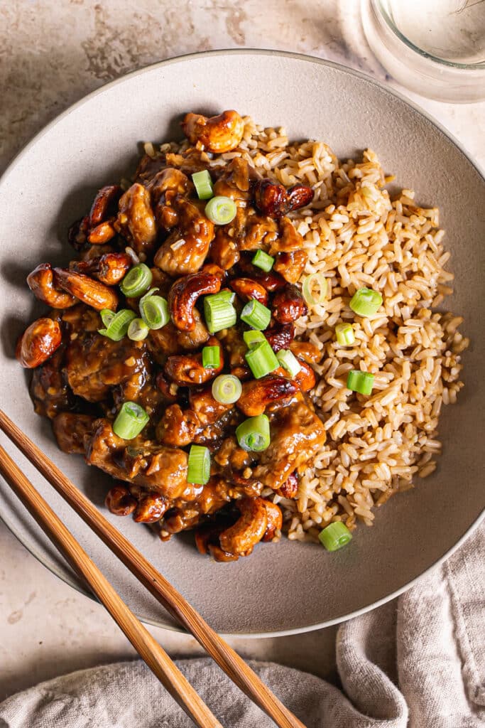 a close up of a bowl of sticky vegan cashew chicken served over brown rice with chopsticks resting on the bowl, a glass of water on the side, a small wooden cutting board with sliced green onions on it.