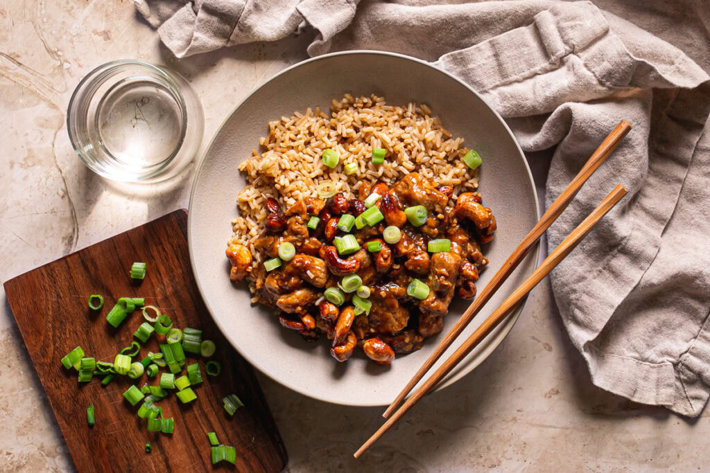 a bowl of sticky vegan cashew chicken served over brown rice with chopsticks resting on the bowl, a glass of water on the side, a small wooden cutting board with sliced green onions on it.