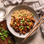a bowl of sticky vegan cashew chicken served over brown rice with chopsticks resting on the bowl, a glass of water on the side, a small wooden cutting board with sliced green onions on it.