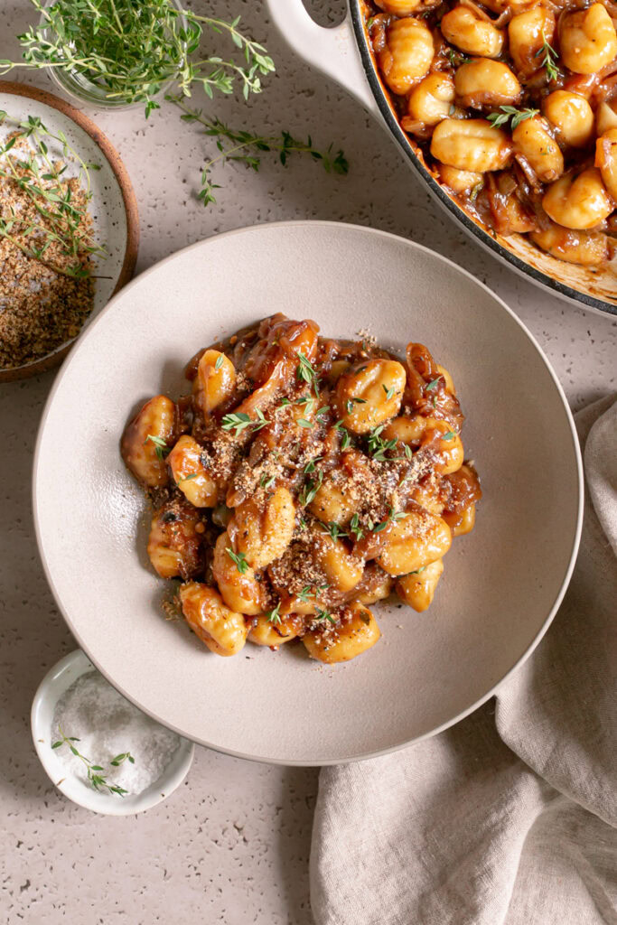 a bowl of French Onion Gnocchi with a small plate of toasted breadcrumbs above it, a small bowl of flake salt below it, a linen napkin, a small glass of fresh thyme leaves and a skillet of French Onion Gnocchi above the bowl.