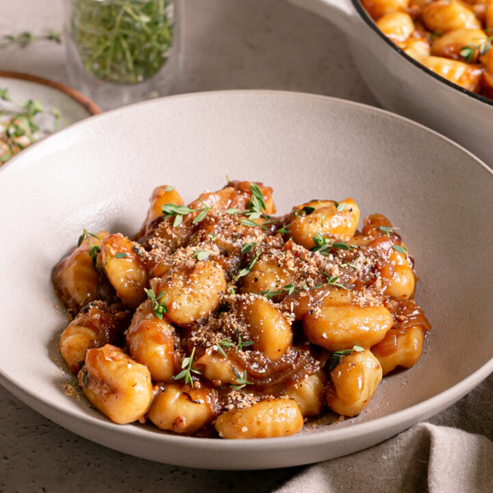 3/4 shot of a bowl of French Onion Gnocchi garnished with fresh thyme leaves and toasted breadcrumbs with the skillet of gnocchi in the background and fresh thyme in a small glass with water