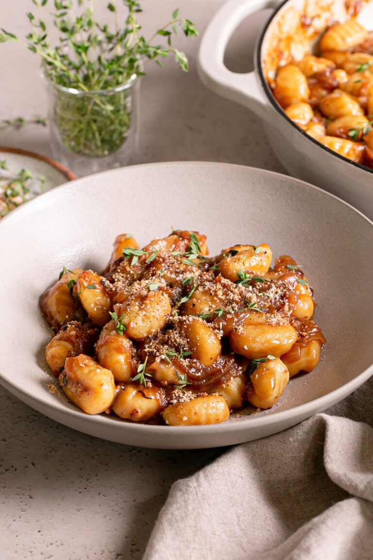 3/4 shot of a bowl of French Onion Gnocchi garnished with fresh thyme leaves and toasted breadcrumbs with the skillet of gnocchi in the background and fresh thyme in a small glass with water