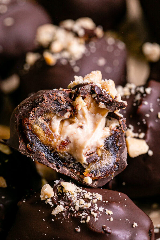 a close up of "Snickers" Frozen Yogurt Chocolate Covered Date Bites on a parchment lined baking sheet, one cut in half to reveal the peanut butter yogurt filling