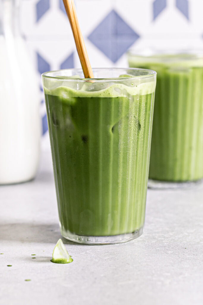 two glasses of iced matcha lattes, one glass with a wooden stir stick sticking out of it and a jug of rice milk in the background