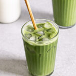 two glasses of iced matcha lattes, one glass with a wooden stir stick sticking out of it and a jug of rice milk in the background