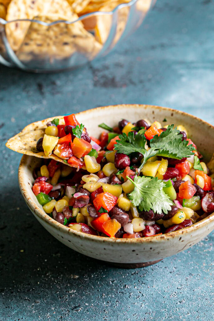 a tortilla chip with cowboy caviar bean dip on it resting on the edge of the bowl with a glass bowl of tortilla chips in the background