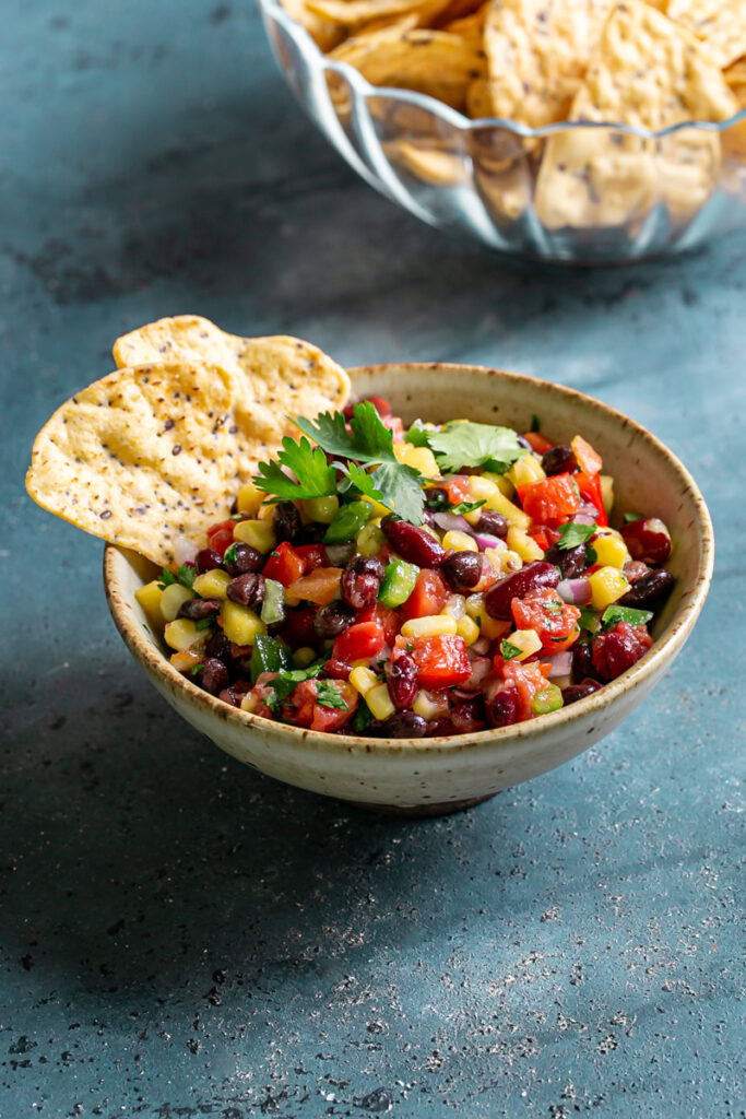 3/4 image of a bowl of cowboy caviar bean dip garnished with fresh cilantro with two tortilla chips sticking out of the bowl and a glass bowl of tortilla chips in the background
