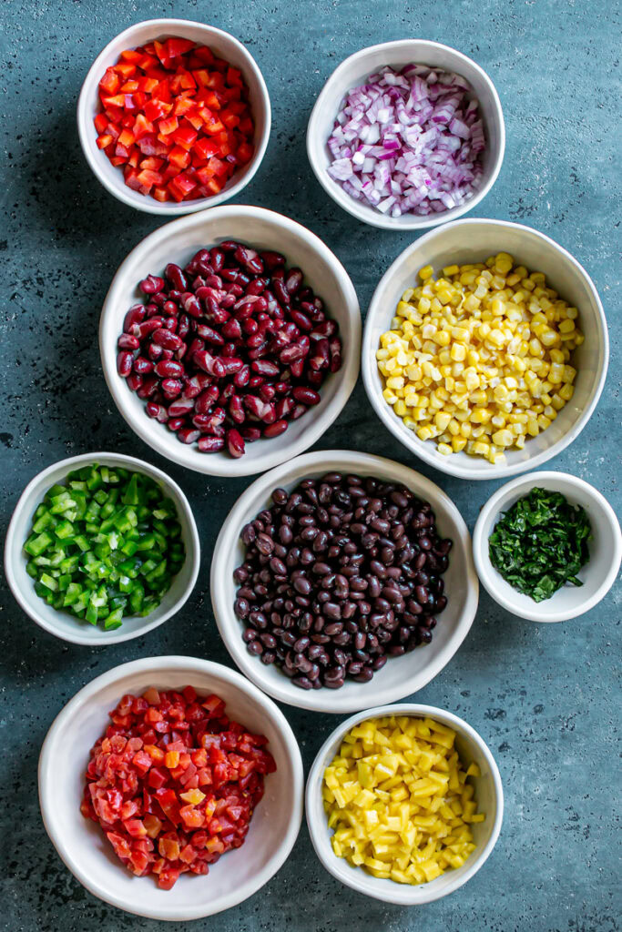 all of the ingredients to make this cowboy caviar dip in separate bowls: diced red and green bell pepper, diced mango, kidney beans, black beans, diced tomatoes, corn, diced red onion, chopped cilantro