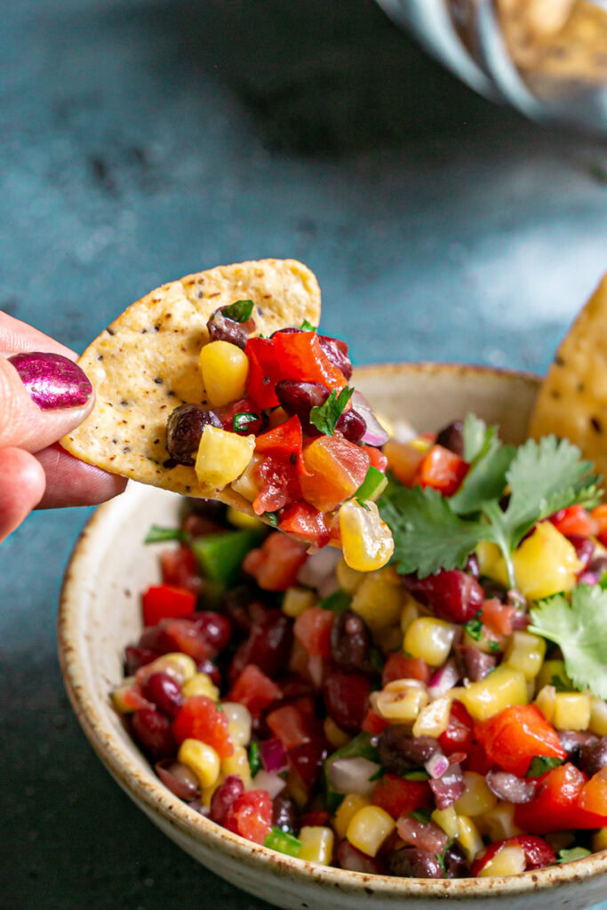 a close up of a hand holding a tortilla chip with cowboy caviar bean dip on it