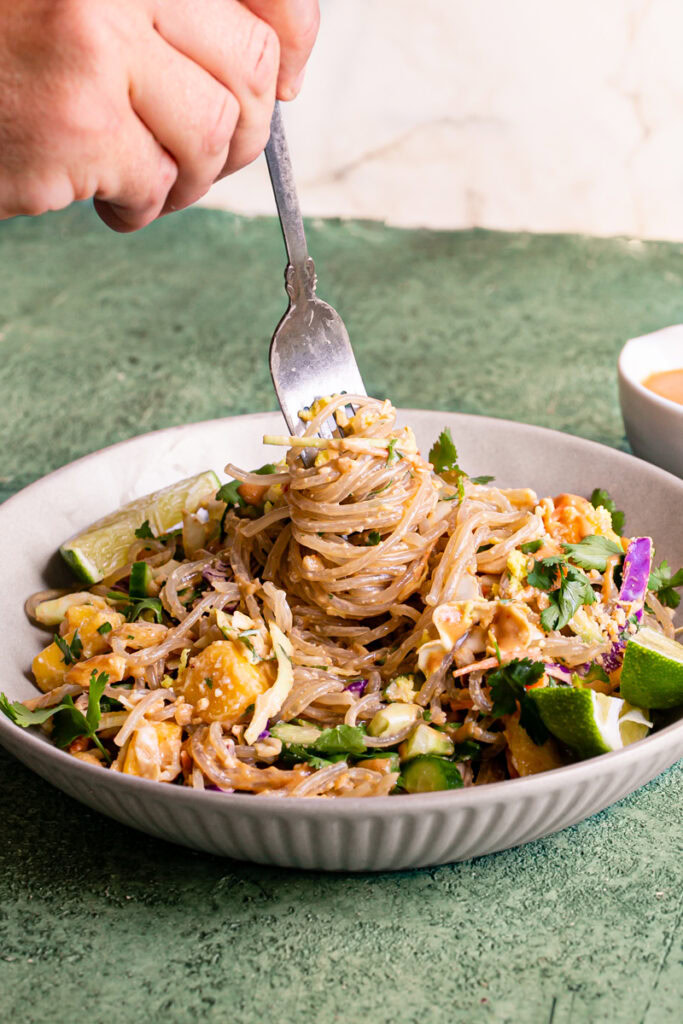 a bowl of Thai Peanut Glass Noodle Salad with lime wedges and a fork in the bowl with noodles. A small bowl of crushed peanuts and a bowl with dressing above it