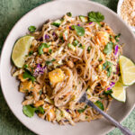 a bowl of Thai Peanut Glass Noodle Salad with lime wedges and a fork in the bowl with noodles spun around it. The Noodles are garnished with fresh cilantro. A small bowl of crushed peanuts and a bowl with dressing above it