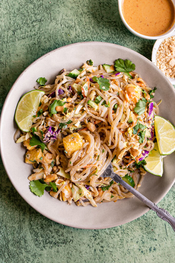 a bowl of Thai Peanut Glass Noodle Salad with lime wedges and a fork in the bowl with noodles spun around it. The Noodles are garnished with fresh cilantro. A small bowl of crushed peanuts and a bowl with dressing above it