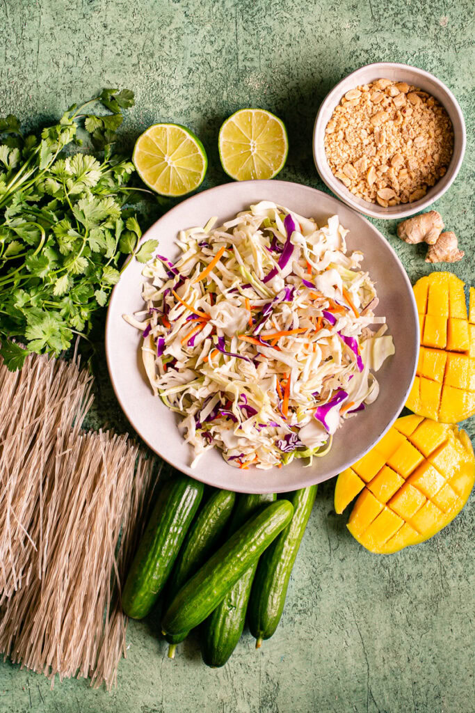 all of the ingredients used to make this Thai Peanut Glass Noodle Salad: dry glass noodles, cucumber, mango, coleslaw mix, fresh cilantro, lime, crushed peanuts, ginger