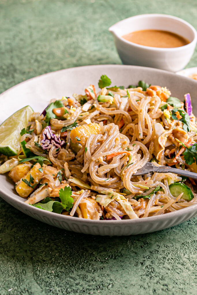 a very close up image of Thai Peanut Glass Noodle Salad in a bowl with a lime wedge and a fork in the bowl with noodles spun around it