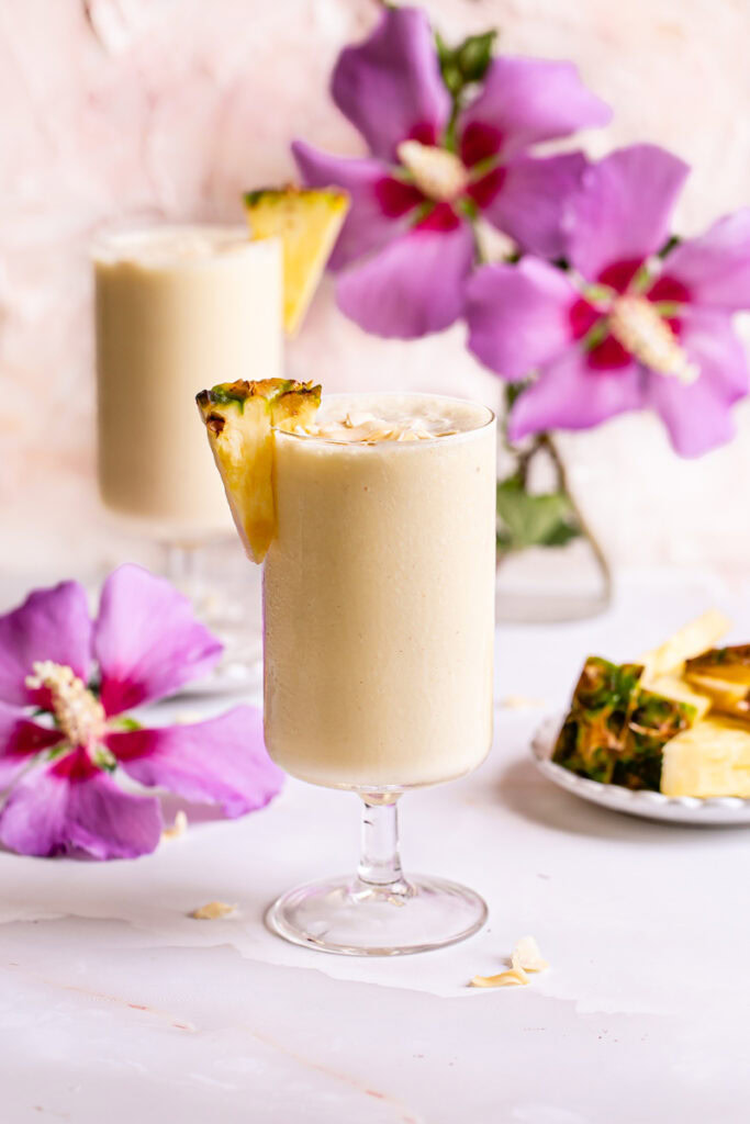 two glasses of 5-Ingredient Pina Colada Smoothie garnished with pineapple slices and coconut chips, a plate of pineapple slices in the background and fresh magenta flowers