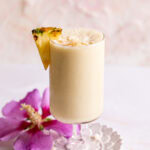 a pina colada smoothie in a glass garnished with a pineapple slice and coconut chips with a magenta flower beside it