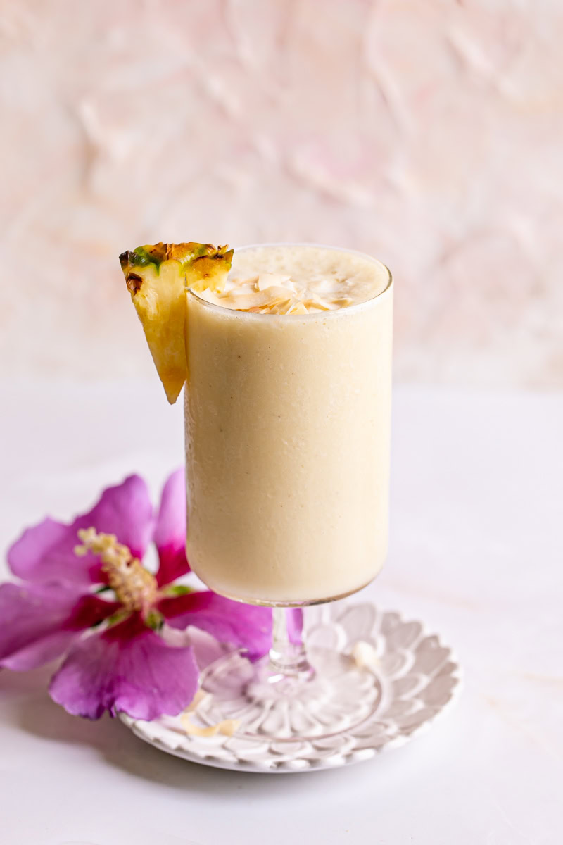a pina colada smoothie in a glass garnished with a pineapple slice and coconut chips with a magenta flower beside it