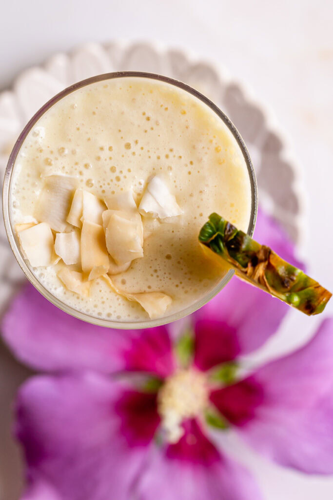 an overhead shot of 5-Ingredient Pina Colada Smoothie garnished with coconut chips and a magenta flower blurred below it