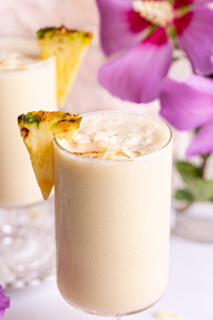 a close up of a glass of 5-Ingredient Pina Colada Smoothie with magenta flowers and another smoothie blurred in the background