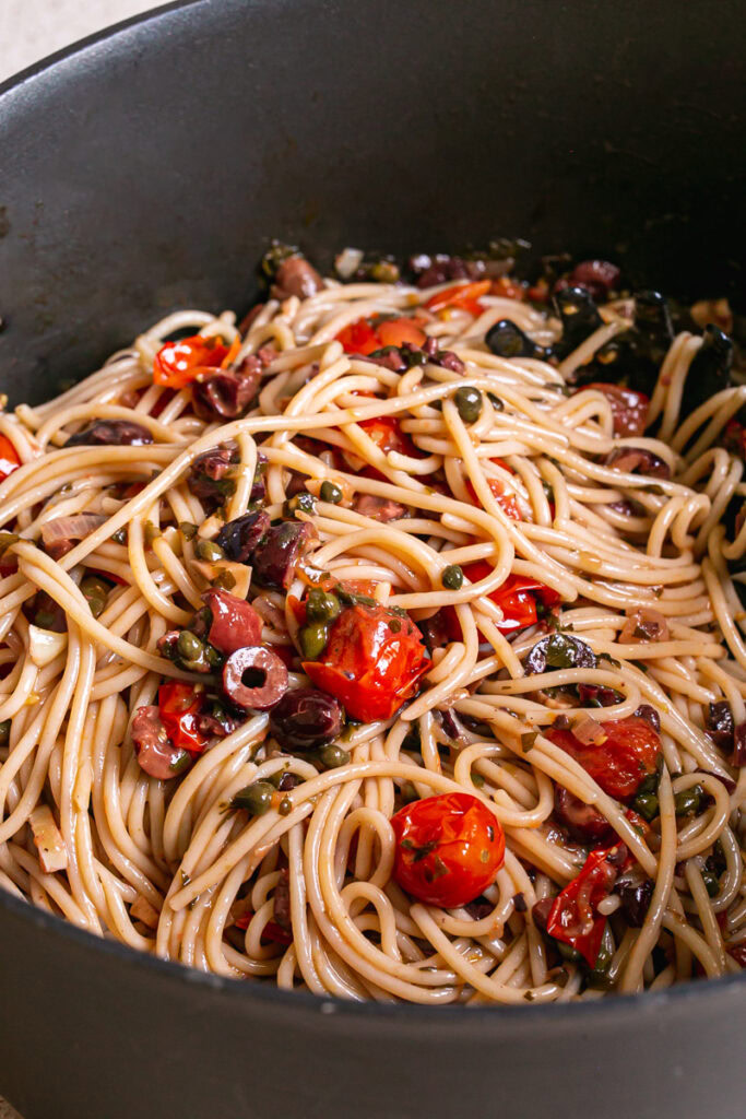 close up 3/4 shot of spaghetti tossed in with the cooked cherry tomato puttanesca sauce ingredients