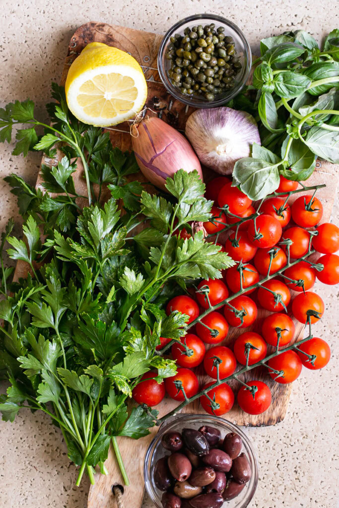 the ingredients used to make Cherry Tomato Puttanesca pasta: fresh parsley, basil, cherry tomatoes, Kalamata olives, capers, shallot, head of garlic, lemon