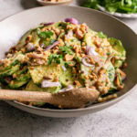3/4 shot of Miso Cucumber Salad with Shallot and Roasted Peanuts with a serving spoon in the bowl and a plate of fresh cilantro and a small bowl of crushed roasted peanuts in the background