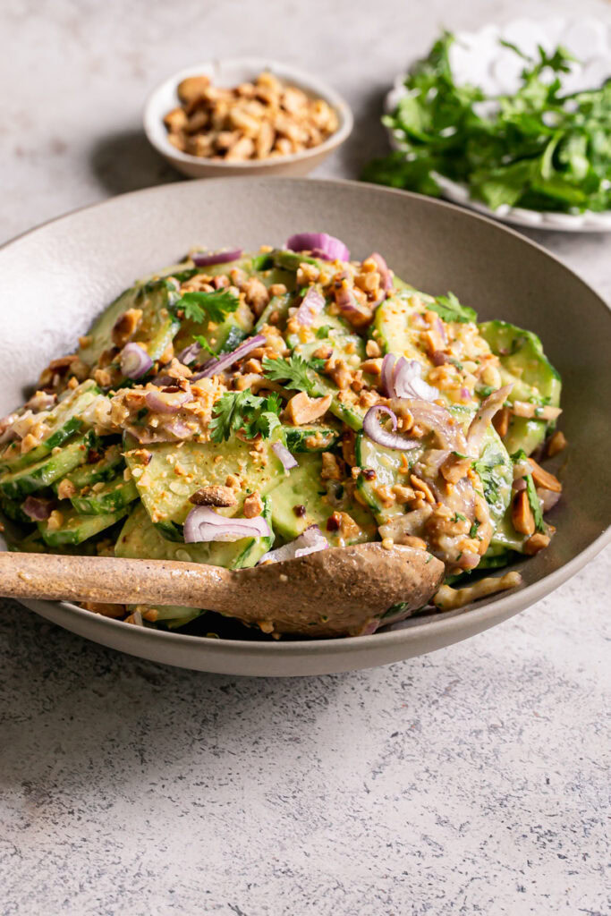 3/4 shot of Miso Cucumber Salad with Shallot and Roasted Peanuts with a serving spoon in the bowl and a plate of fresh cilantro and a small bowl of crushed roasted peanuts in the background