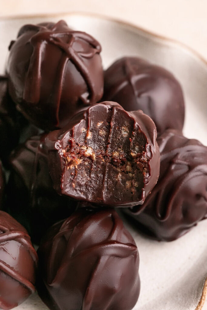 a close up of a plate of chocolate crunch date truffles piled on top of one another, one with a bite taken out of it