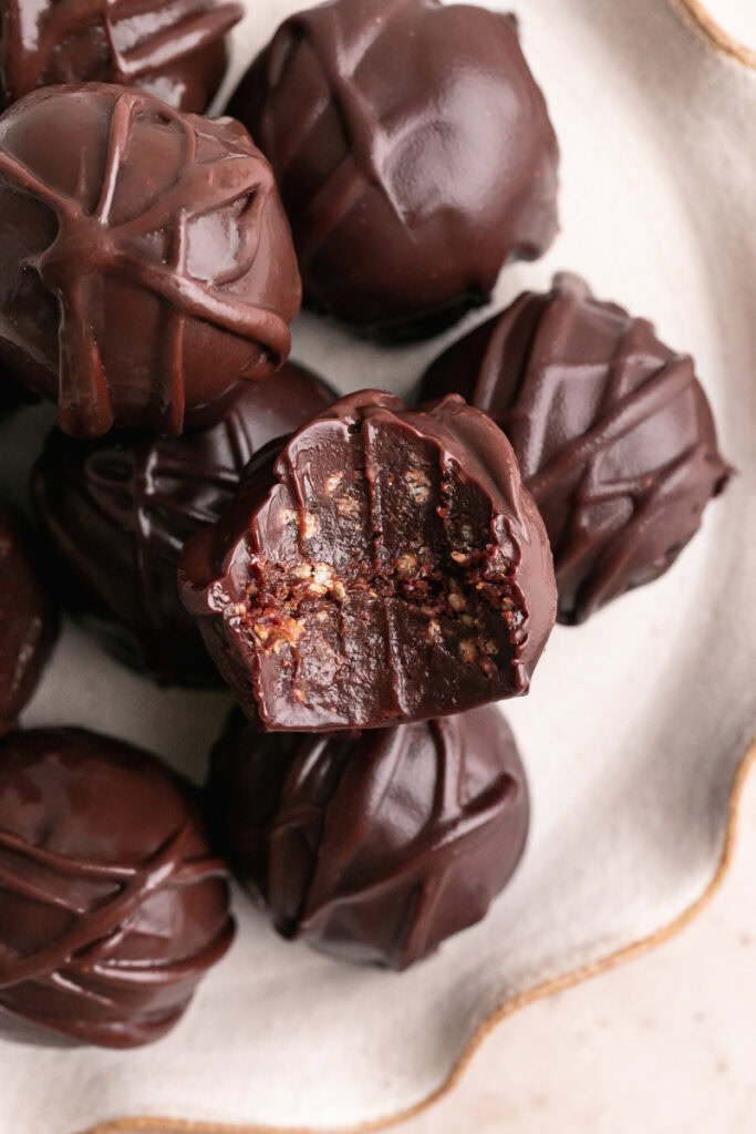 a close up of a plate of chocolate crunch date truffles piled on top of one another, one with a bite taken out of it