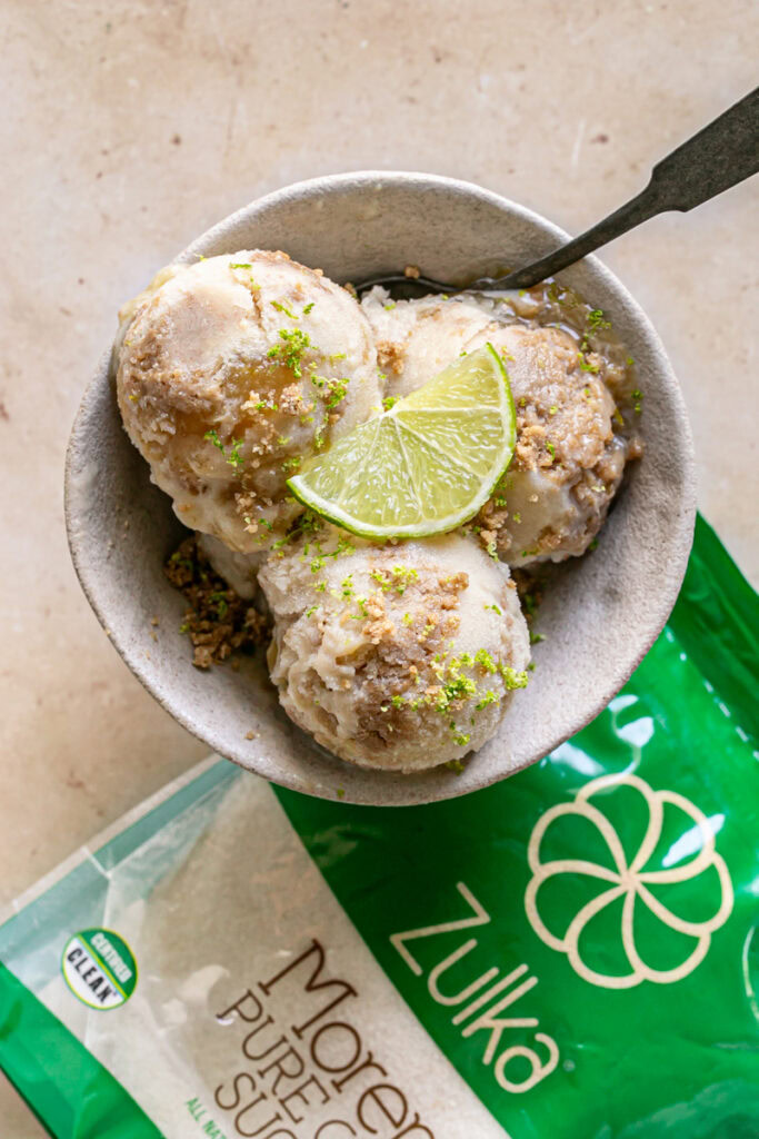 three scoops of No-Churn Key Lime Pie Ice Cream in a bowl garnished with a slice of lime, cookie dough crumble and lime zest with a bag of Zulka morena pure cane sugar next to the bowl