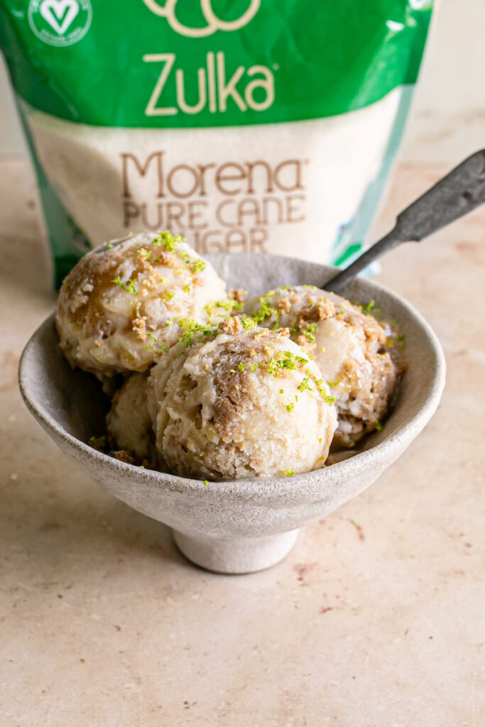 a bowl filled with 3 scoops of No-Churn Key Lime Pie Ice Cream with a spoon in it and a bag of Zulka morena pure cane sugar in the background