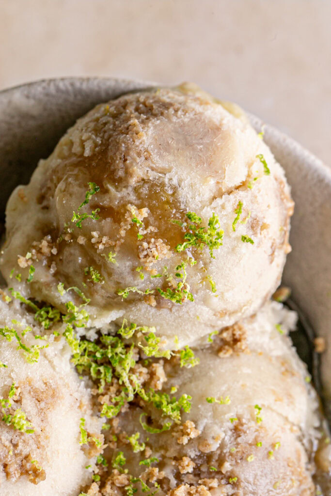 a close up of a scoop of No-Churn Key Lime Pie Ice Cream in a bowl with 2 other scoops garnished with cookie dough crumble and lime zest