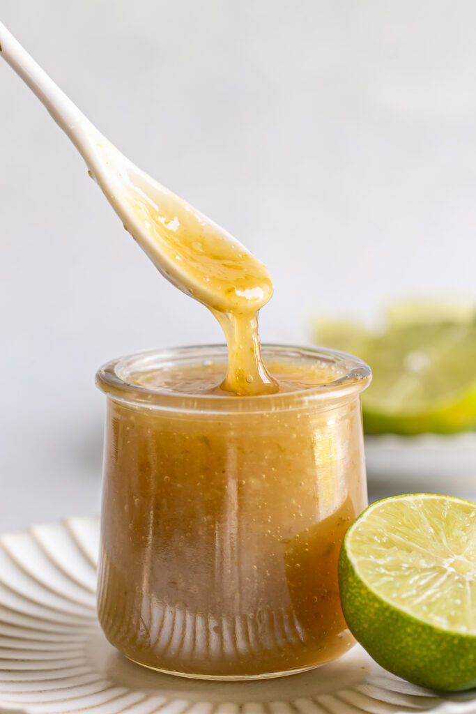 vegan lime curd in a small glass jar with a spoon scooping some out and limes in the background