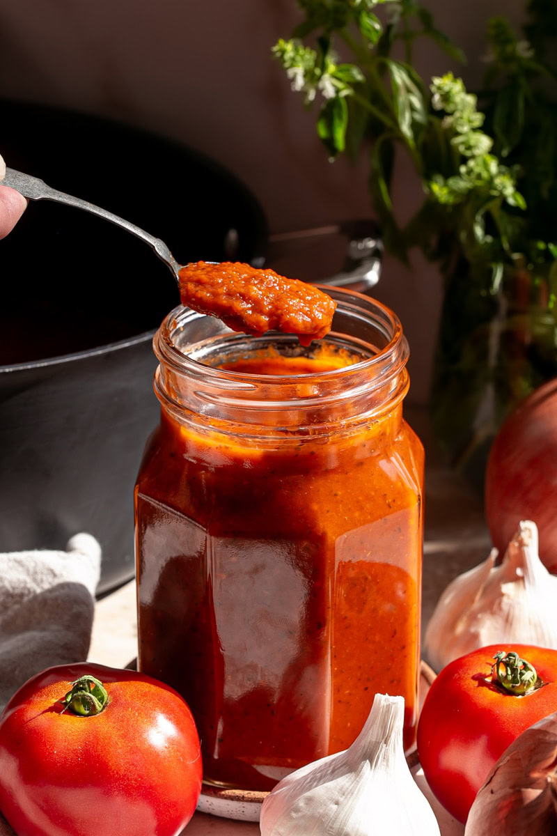 a jar of Caramelized Onion and Garlic Roasted Tomato Sauce with a spoonful of sauce coming out of the jar, surrounded by fresh tomatoes, onion, basil and garlic