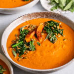 3/4 shot of a bowl of Roasted Butternut Squash Tomato Soup garnished with with Crispy Kale and toasted pumpkin seeds and fresh sage leaves, another bowl in the background