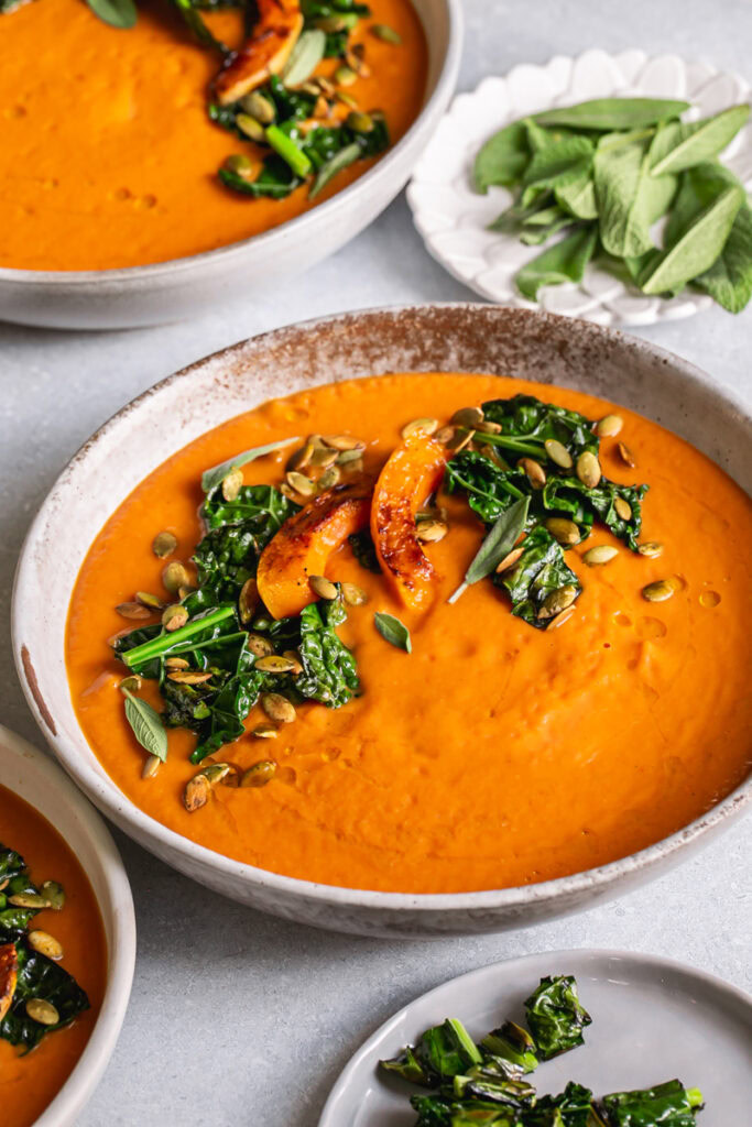 3/4 shot of a bowl of Roasted Butternut Squash Tomato Soup garnished with with Crispy Kale and toasted pumpkin seeds and fresh sage leaves, another bowl in the background