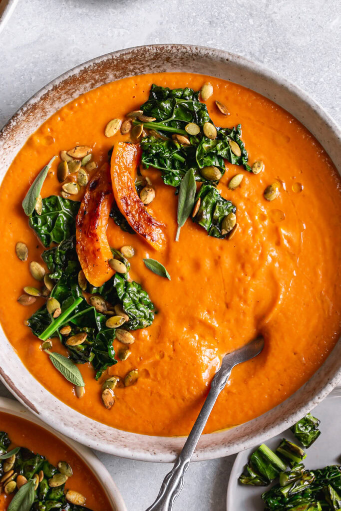 a close up of a bowl of three bowls of Roasted Butternut Squash Tomato Soup garnished with with Crispy Kale and toasted pumpkin seeds and fresh sage leaves with a spoon in the bowl