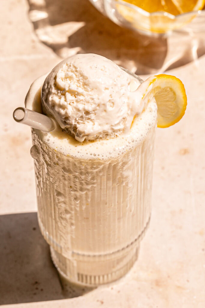 3/4 shot of an overflowing glass of lemonade milkshake with a scoop of vanilla ice cream on top, a glass straw sticking out and a slice of lemon on the glass with a blow of lemons in the background