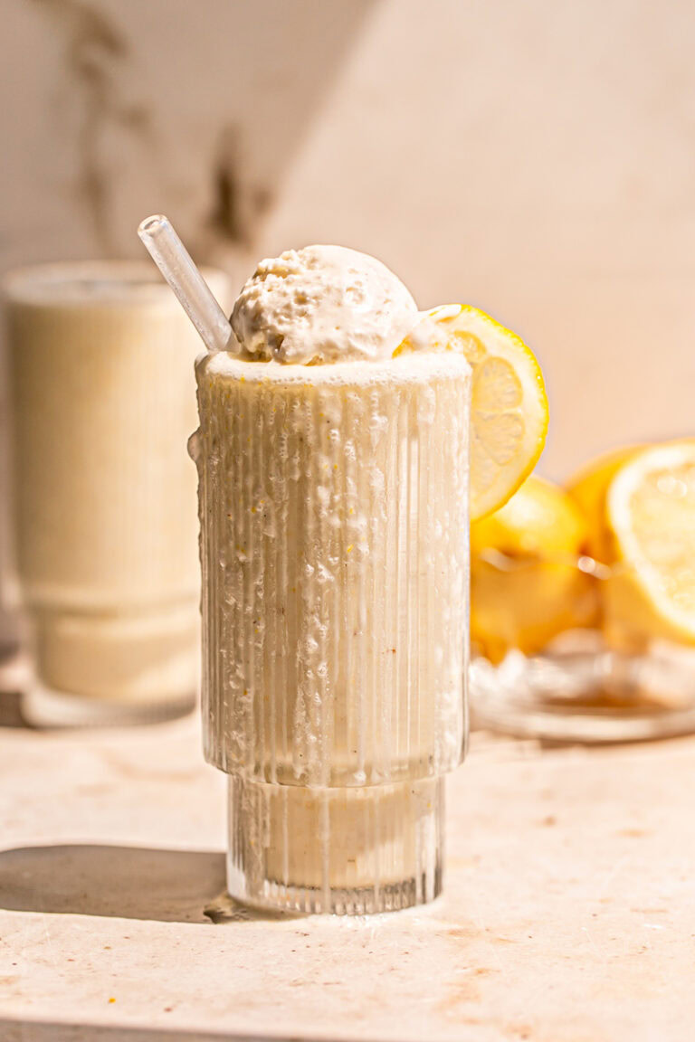 two glasses of lemonade milkshake, one overflowing down the glass topped with a scoop of vanilla ice cream and a bowl of lemons in the background