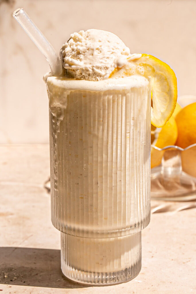 an overflowing glass of lemonade milkshake with a scoop of vanilla ice cream on top, a glass straw sticking out and a slice of lemon on the glass