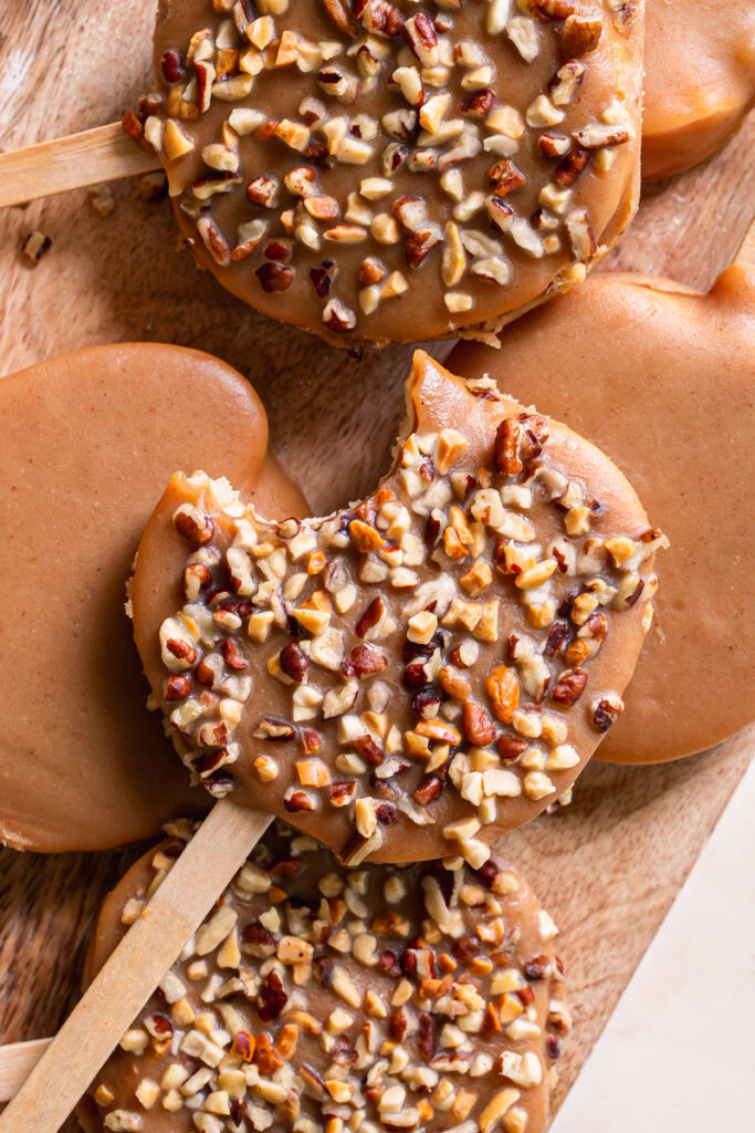 caramel apple slices on popsicle sticks, some garnished with chopped nuts and others not, the top one with a bite taken out of it
