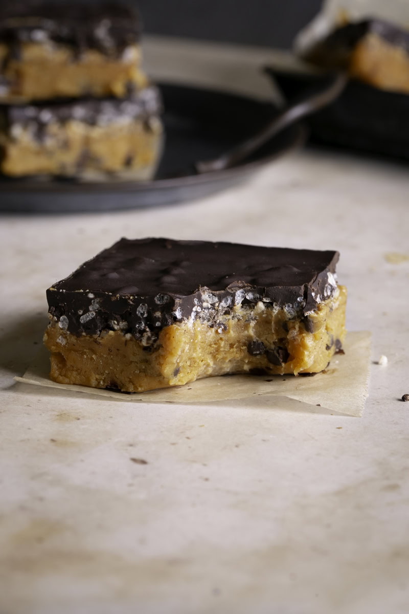one Almond Flour Pumpkin Cookie Dough Crunch Bar on a piece of parchment paper, a bite taken out of it, and more bars in the background