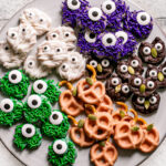 a round plate with chocolate covered one eyed green and purple sprinkle covered pretzel monsters, white chocolate covered pretzel mummies, orange pumpkin pretzels, and chocolate covered owl pretzels