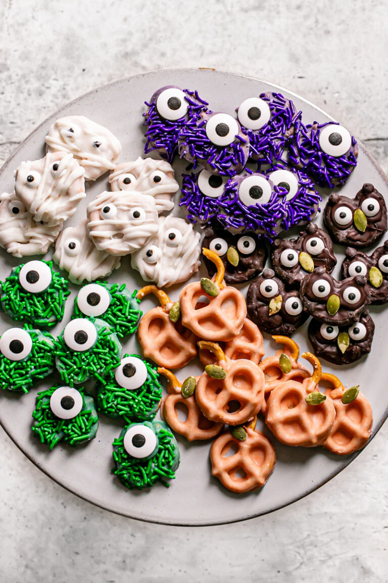 a round plate with chocolate covered one eyed green and purple sprinkle covered pretzel monsters, white chocolate covered pretzel mummies, orange pumpkin pretzels, and chocolate covered owl pretzels