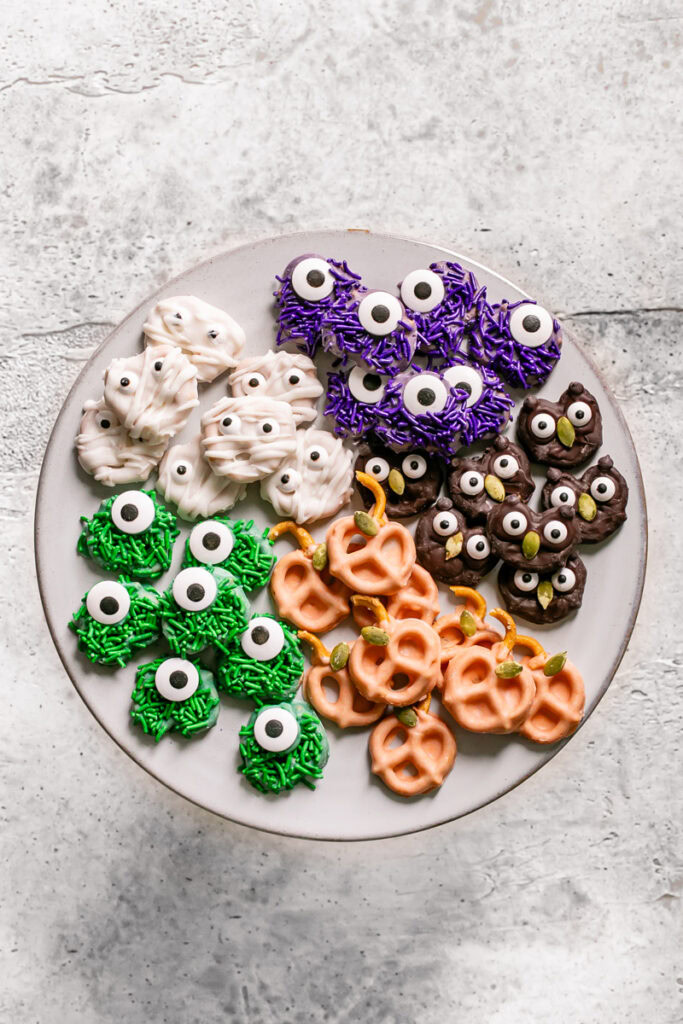 a round plate with chocolate covered one eyed green and purple sprinkle covered pretzel monsters, white chocolate covered pretzel mummies, orange pumpkin pretzels, and chocolate covered owl pretzels