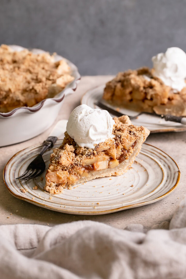 a slice of healthy dutch apple pie with a scoop of vanilla ice cream on top and the rest of the pie in the pie plate in the background